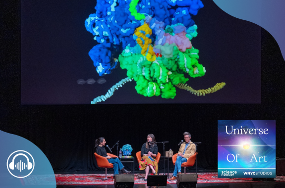 A large colorful 3-d model of a complex biological subject is projected above three people speaking on a stage.