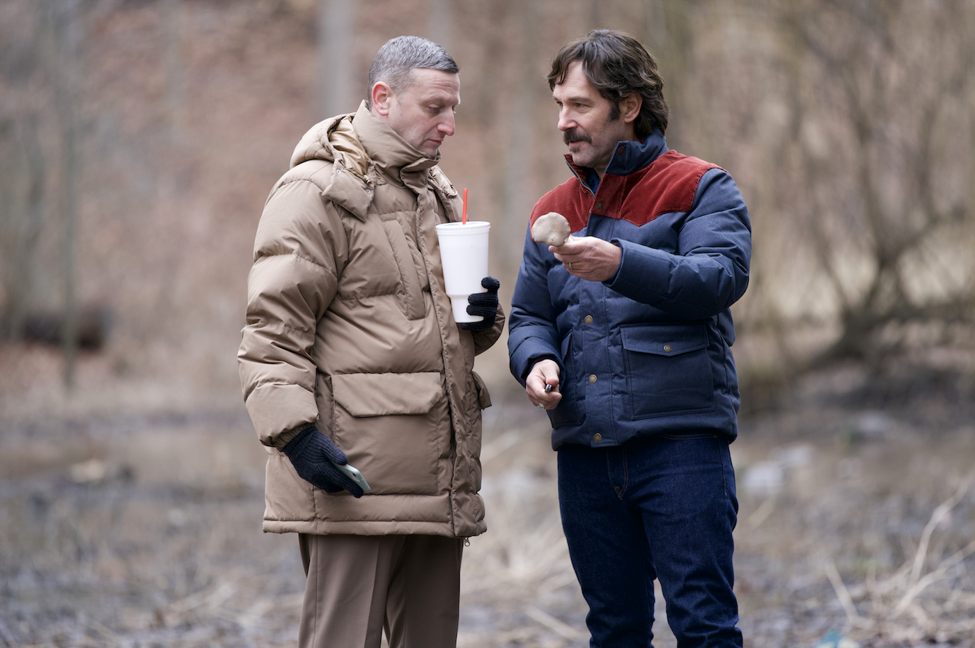 Two men wearing warm outerwear stand in a forest. One holds a soda and one holds a mushroom.