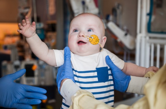 A baby with a tube up his nose looking happy and healthy, as a pair of gloved hands holds him