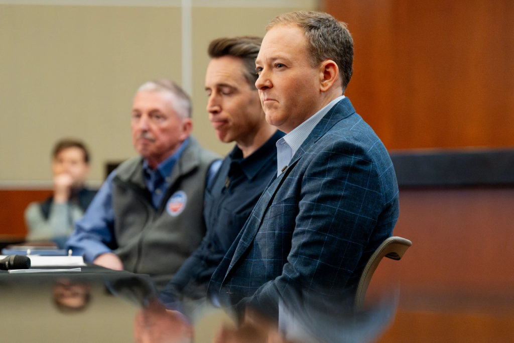 Three white men in blue suits and shirts sit along a table in a wood-paneled room looking to the left side of the frame.