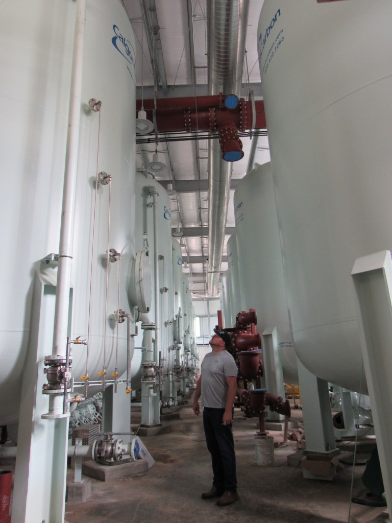 A man in dark pants and a t-shirt stands between rows of massive white tanks, looking up.
