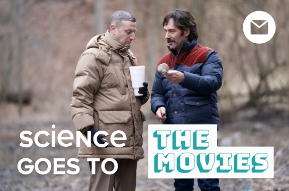 Two men wearing warm outerwear stand in a forest. One holds a soda and one holds a mushroom.