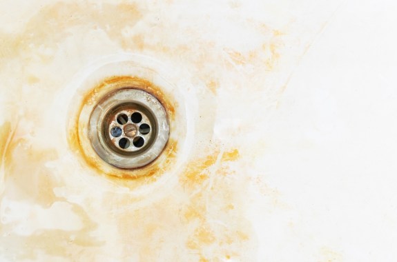 An old rusty bathtub surface with a metal drain hole. Dirty cracked unclean bath or sink with red microbe goo stain, close-up. Corrosion, unsanitary concept, copy space.