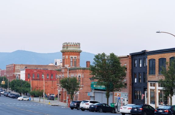 A broad street lined by brick apartment buildings and businesses, with low mountains in the background.