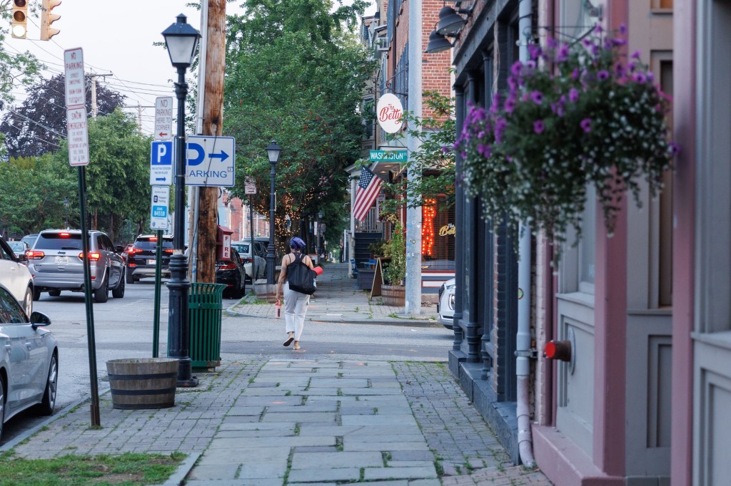 Looking down a sidewalk of a small town with storefronts on the right and cars on the left. One person with a black bag walks away down the sidewalk.