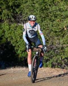 A man on a bike smiles as he pedals toward the camera, with greenery in the background.