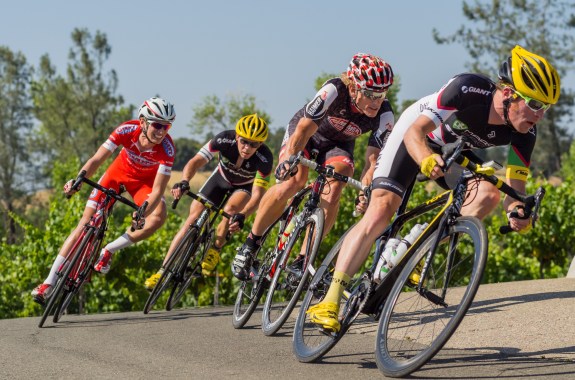 4 cyclists lean to the right as they round a curve on a sunny day