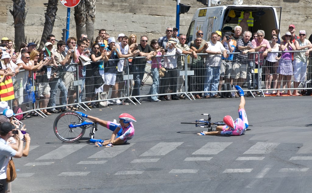 Two cyclists in pink uniforms are sprawled on black asphalt while fans look on.