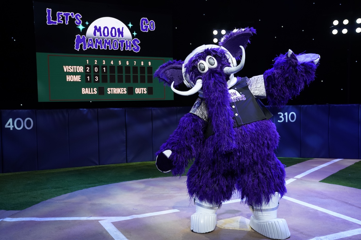 A purple mammoth mascot wearing an astronaut helmet. It's standing on a set of a baseball field with a sign that says "let's go Moon Mammoths"
