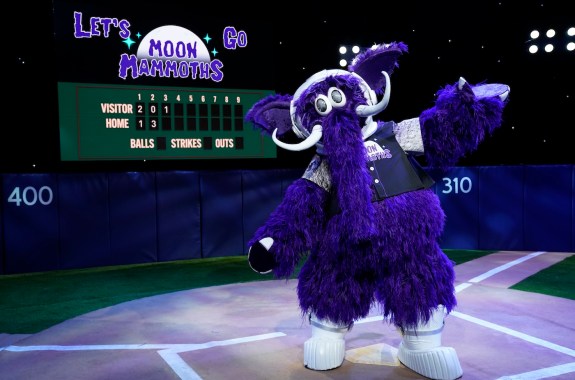A purple mammoth mascot wearing an astronaut helmet. It's standing on a set of a baseball field with a sign that says "let's go Moon Mammoths"