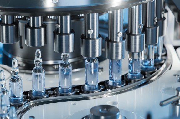 Close-up of small vials in a line being filled with clear liquid through metal tubes on a machine.