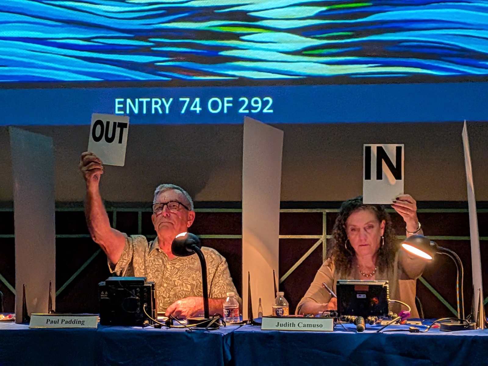 Two people each hold up a card. The person on the left holds a card that reads "OUT" and the person on the right holds up a card that reads "IN."