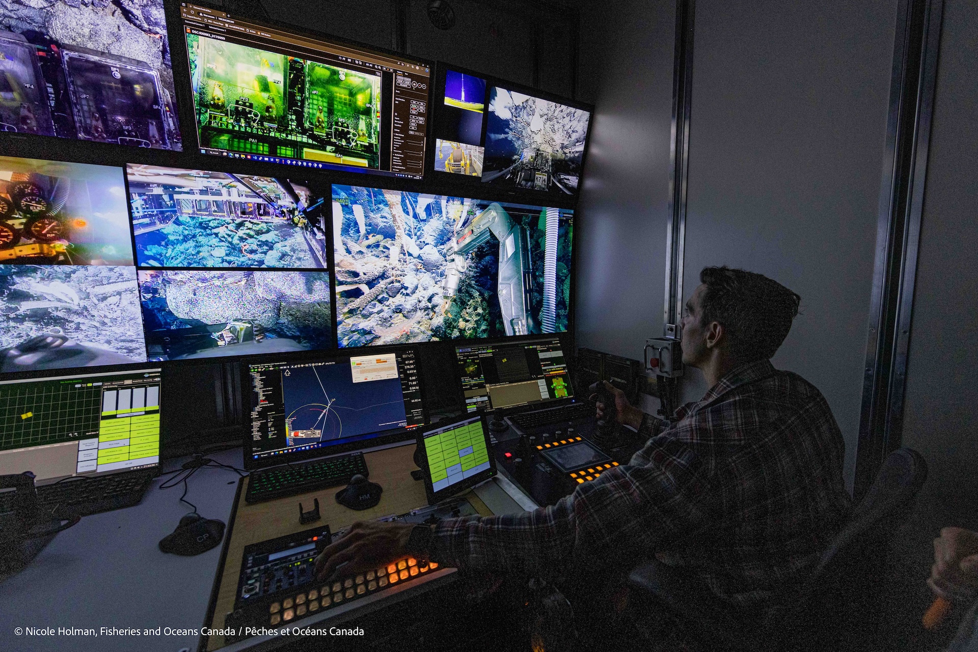 A person sits in a dimly lit area looking at several monitors showing colorful scenes from the ocean floor.