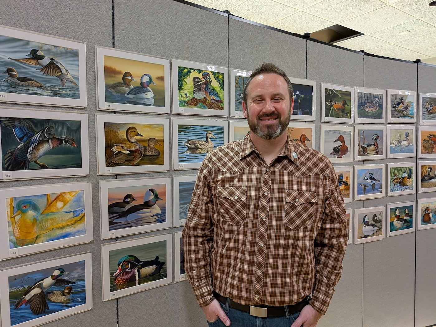 A man stands in front of 4 rows of paintings of ducks. He's at the 2025 Federal Duck Stamp Art Contest
