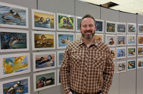 A man stands in front of 4 rows of paintings of ducks. He's at the 2025 Federal Duck Stamp Art Contest
