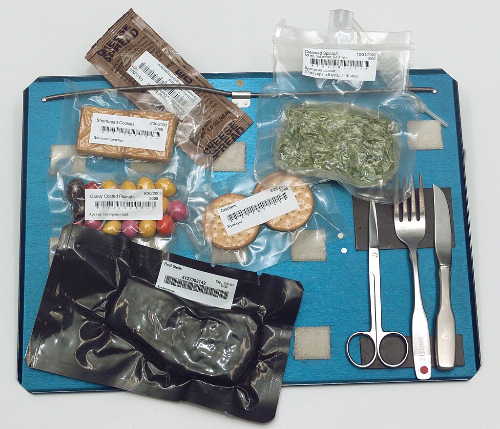 Space food meal laid out on blue tray with various vacuum-sealed packages including candy-coated peanuts, crackers, creamed spinach, shortbread cookies, and beef steak, alongside metal utensils (knife, fork, scissors).