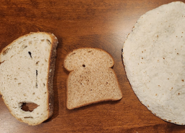 Three foods on a wooden cutting board: a slice of crusty sourdough with holes, a slice of whole-wheat bread, and a large flour tortilla.