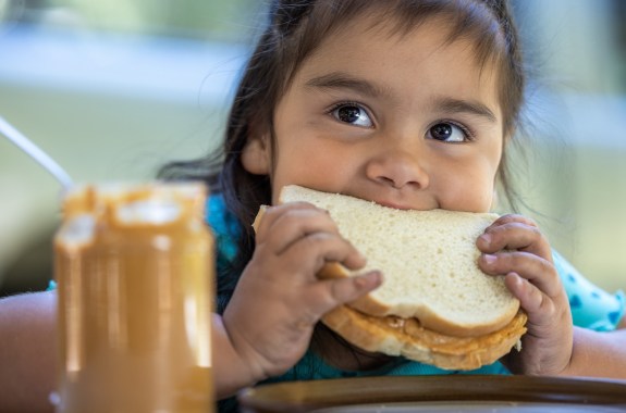 A young girl is eating a sandwich with a jar of peanut butter next to her. The scene is casual and relaxed, with the girl enjoying her snack. peanut allergies prevention